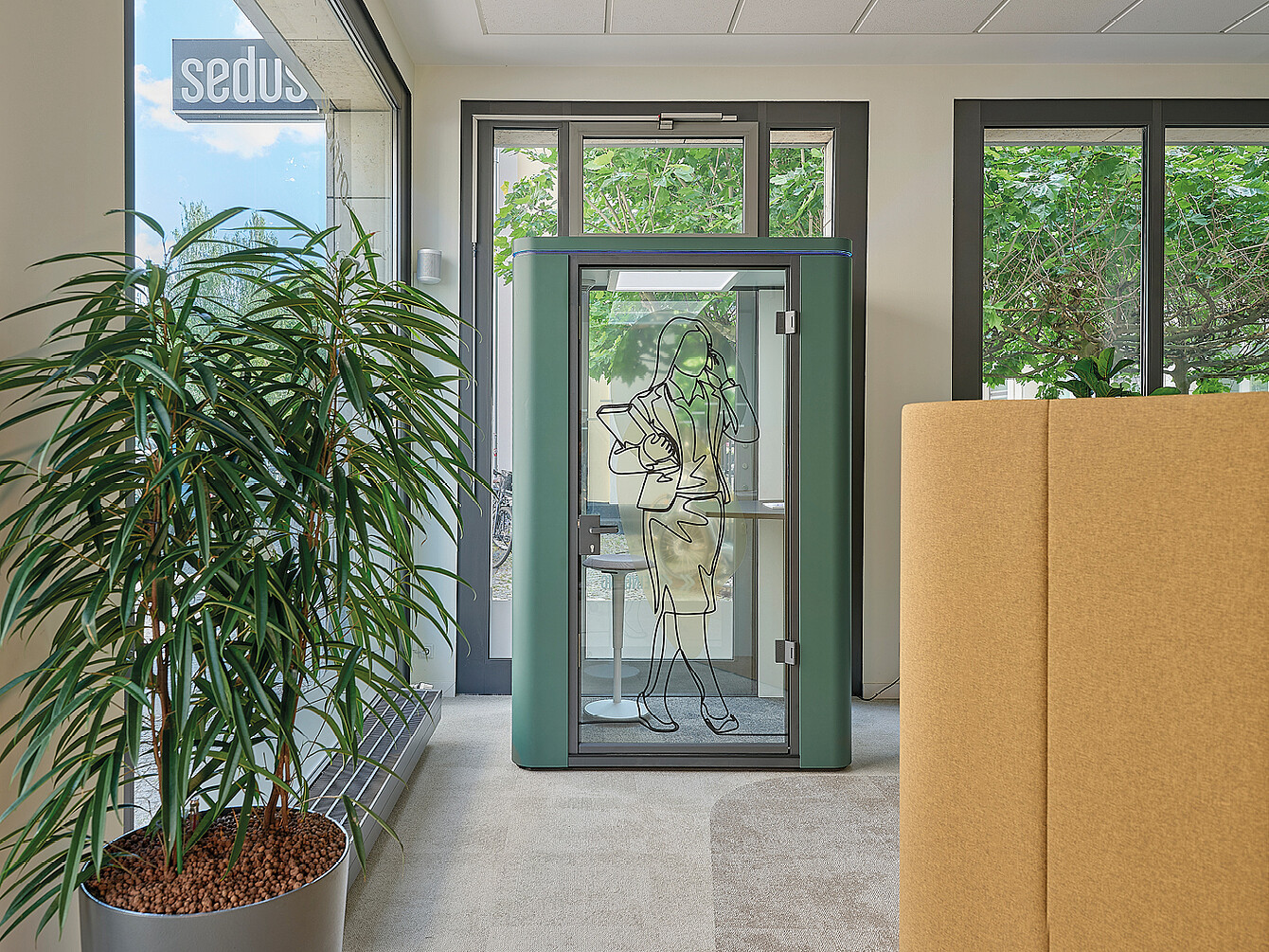 Petite cabine insonorisée verte avec une porte vitrée sur laquelle est dessiné une femme tenant un téléphone, placée dans un espace de bureau moderne près de grandes fenêtres et d’une grande plante en pot.