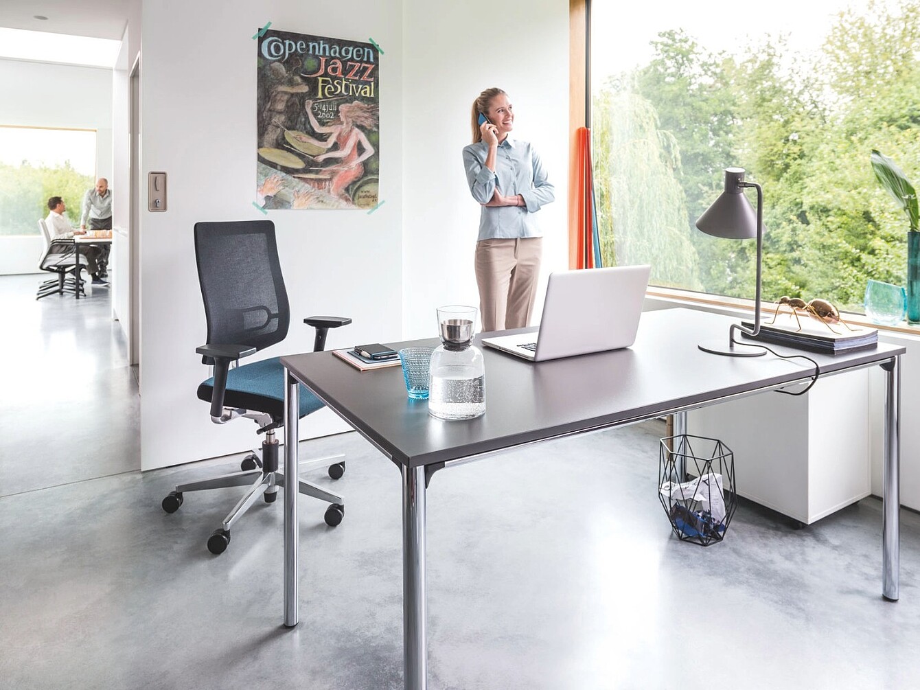 Bureau moderne avec un siège ergonomique en résille noire, un bureau sur lequel se trouvent un ordinateur portable, une bouteille d’eau en verre, une lampe, et une femme debout près d’une grande fenêtre en train de parler au téléphone