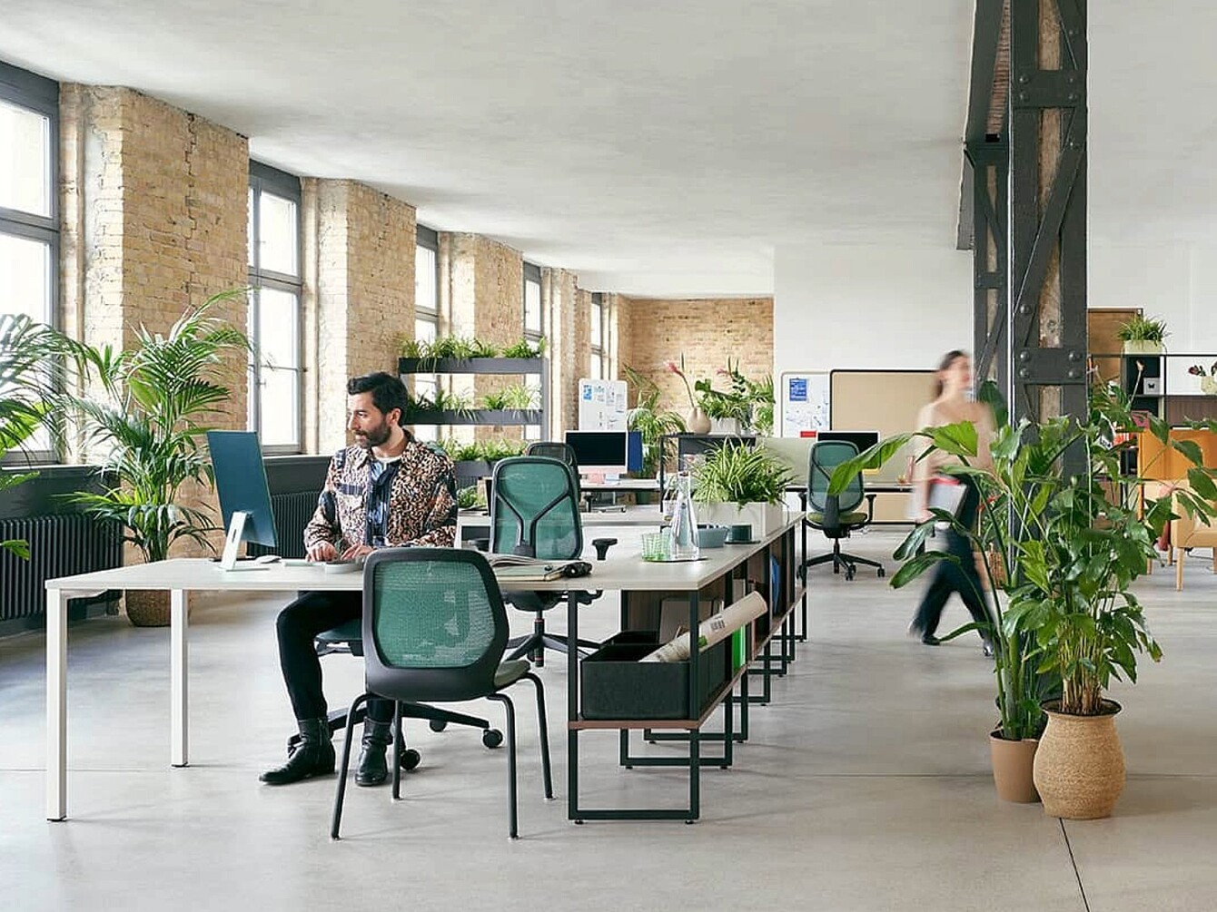Paisaje de oficina abierto y luminoso con varios escritorios, sillas de oficina ergonómicas, estanterías y numerosas plantas.