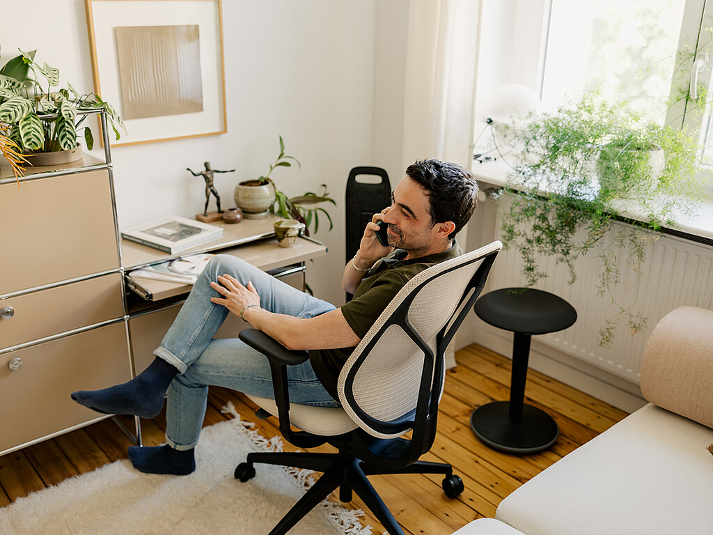 Person sitzt auf ergonomischem Bürostuhl mit Netzrückenlehne, telefoniert im hellen Homeoffice mit Pflanzen und Holzboden