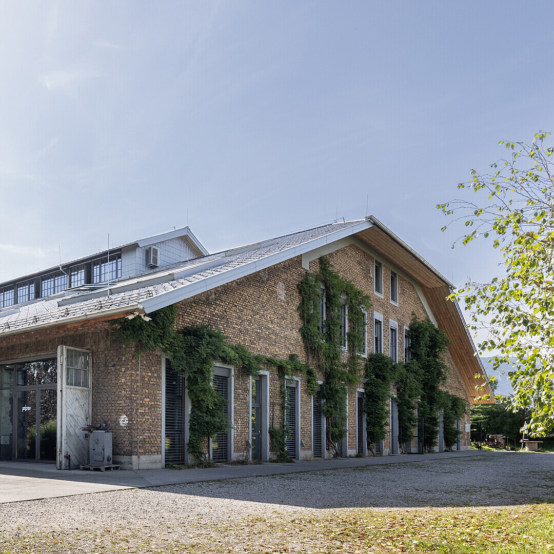 Low-rise brick building with large windows and a sloped metal roof surrounded by greenery under a clear sky