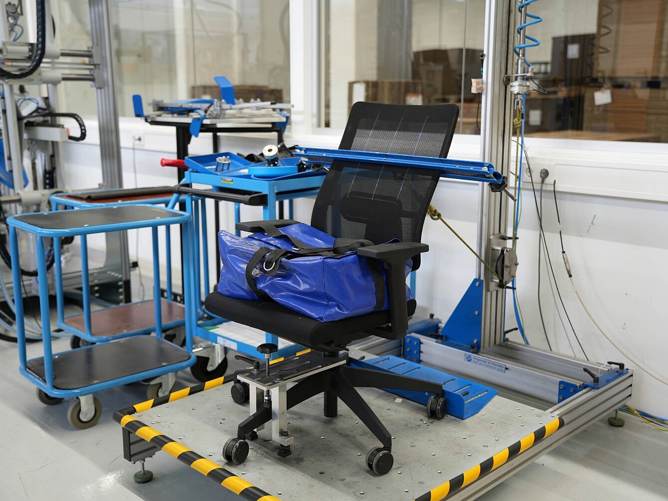 Bureaustoel vastgezet in een blauw metalen testframe in een laboratorium, met apparatuur en karren op de achtergrond
