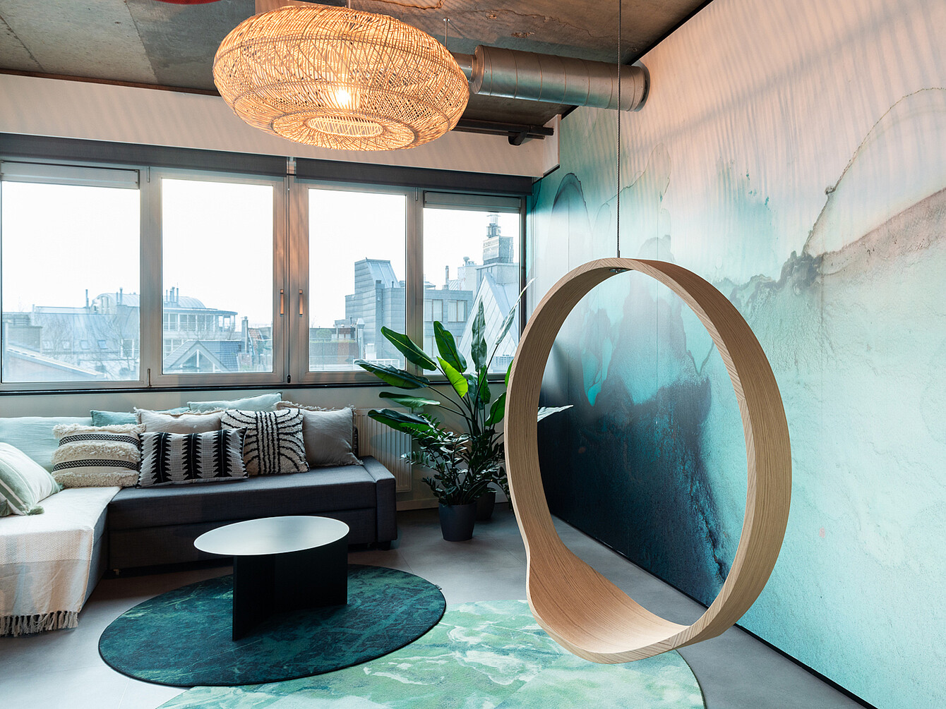 Salon de bureau moderne avec un canapé modulable gris, des coussins à motifs, une table basse ronde noire posée sur un tapis vert, une grande plante, une chaise suspendue circulaire en bois et une grande fenêtre donnant sur les immeubles de la ville