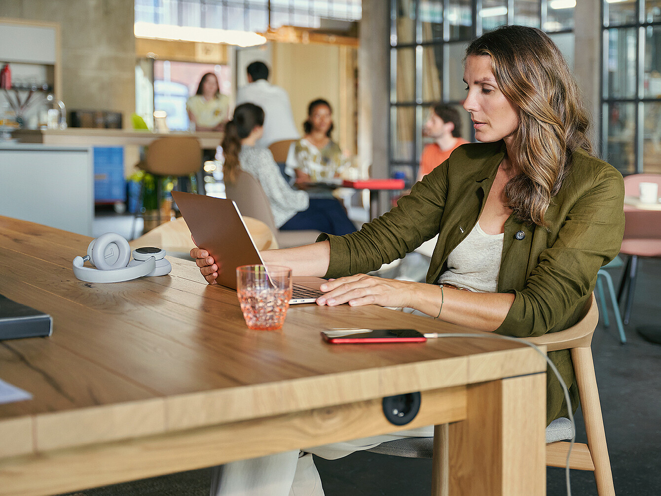 Femme assise à une table en bois dans un espace de travail Work Café, utilisant une tablette avec des écouteurs et un verre à proximité, d’autres personnes discutant en arrière-plan