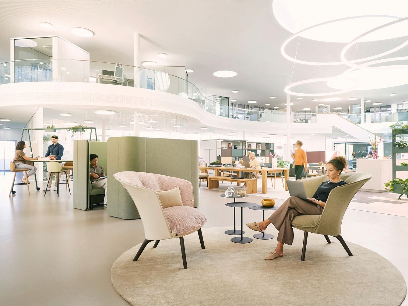 Espacio de oficina abierto y moderno con una mujer sentada en un sillón beige leyendo un libro, una pequeña mesa redonda a su lado y otras personas trabajando o conversando al fondo