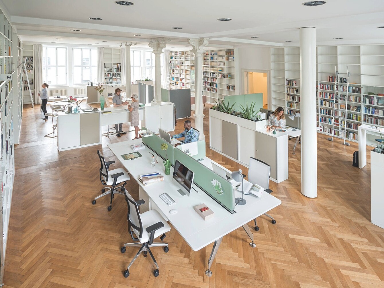 Open kantoorruimte met bureaus gescheiden door groene stoffen panelen, meerdere mensen aan het werk, boekenkasten langs de muren en houten parketvloer