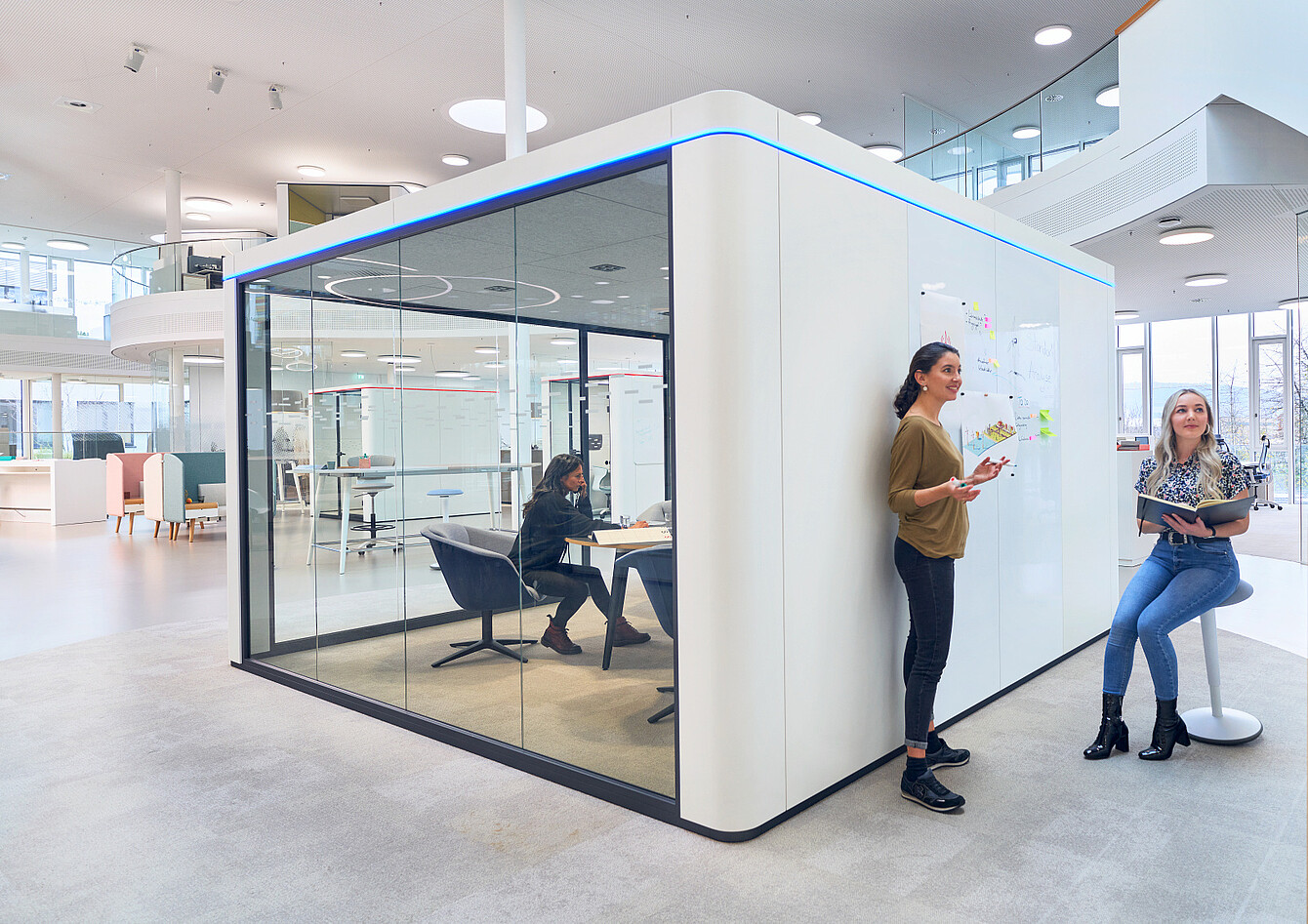 Espacio de oficina moderno con una sala de reuniones con paredes de cristal y dos mujeres de pie y sentadas junto a una pared con pizarra blanca y notas