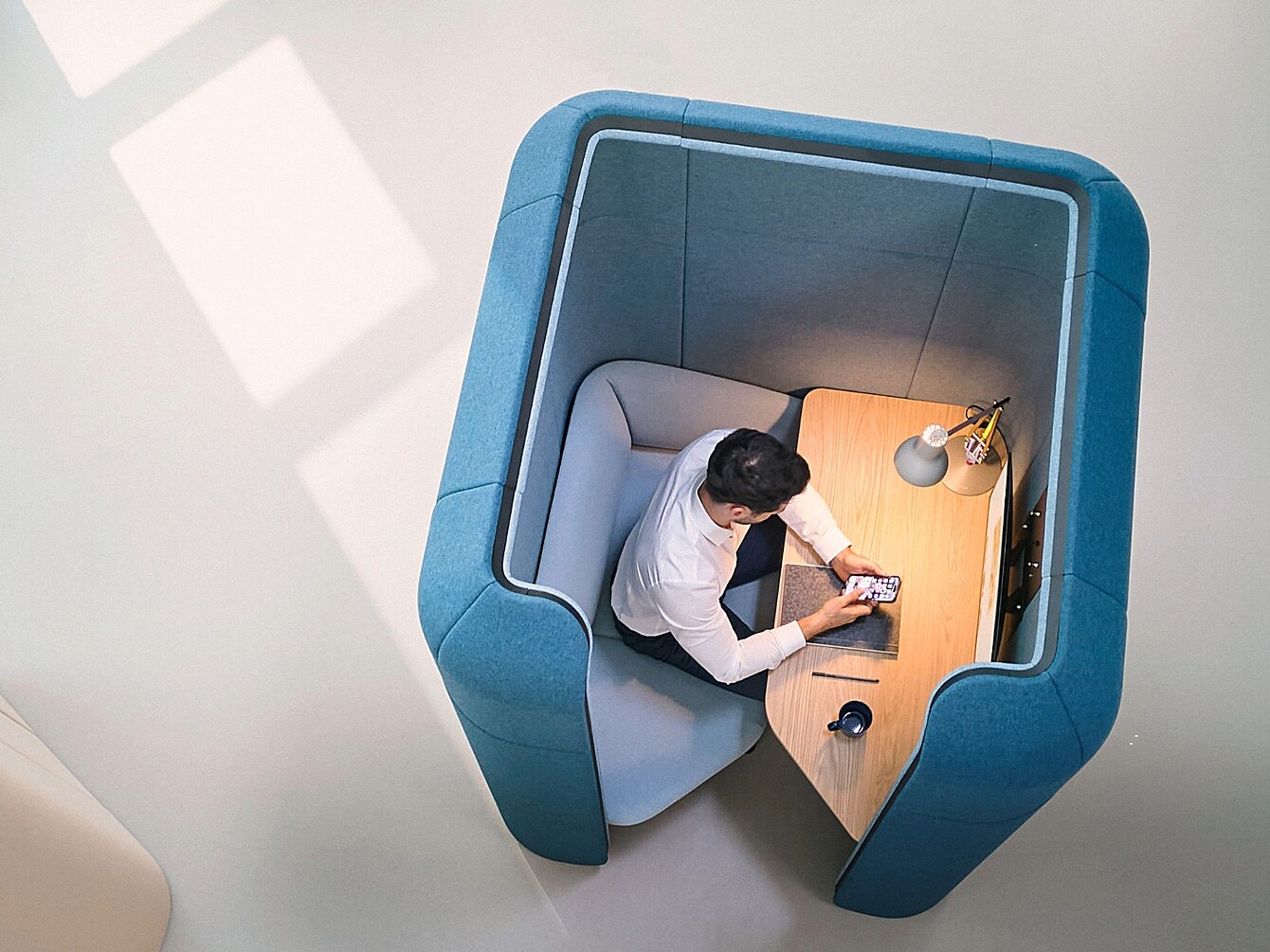 Vue de dessus d’une personne assise à l’intérieur d’une capsule modulaire se:hive aux parois capitonnées, utilisant un smartphone et un ordinateur portable sur un petit bureau en bois.
