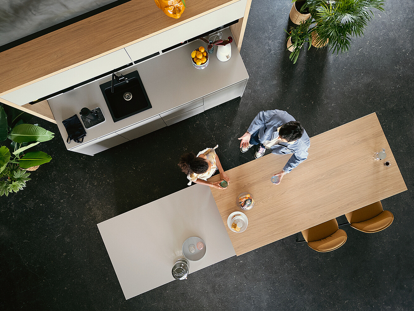 Zwei Personen an einem langen Holztisch in einer modernen, hybriden duktile Gemeinfläche mit Küchenzeile und Pflanzen, die Flexibilität und Gemeinschaft im Arbeitsumfeld symbolisiert