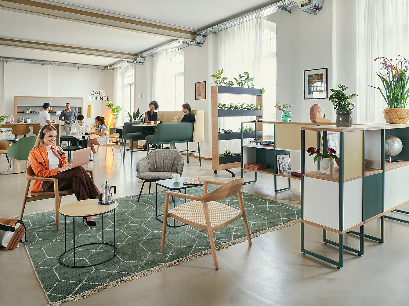 Een modern Work Café met verschillende zitjes met planten. Op de voorgrond zit een vrouw in een oranje jas op een stoel met een laptop. Op de achtergrond zijn mensen in gesprek en aan het werk aan tafels. De ruimte bevat planken met decoratieve voorwerpen en een bordje met 'CAFE LOUNGE'.