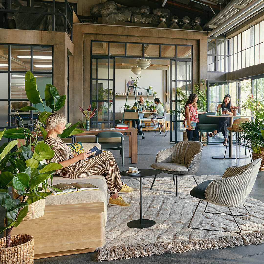 Offenes Büro mit großen Fenstern, vielen Pflanzen und modernen Sitzmöbeln, die eine naturnahe Arbeitsumgebung im Work Café Archetyp Garten schaffen