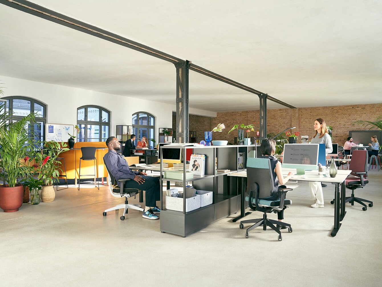 Large open-plan office with several people at desks, plants, and exposed brick wall in the background