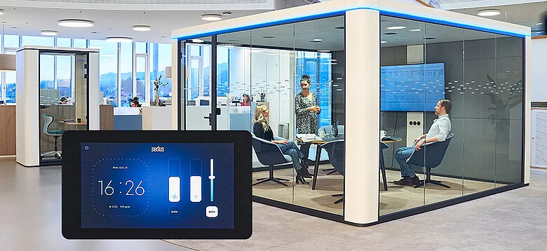 Espace de bureau moderne avec une salle de réunion aux parois vitrées où trois personnes sont assises et une personne est debout, avec un écran tactile numérique affichant l’heure et les commandes au premier plan.